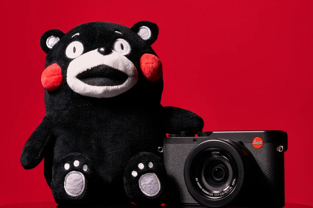 Black Kumamon plush beside a black Leica camera against a solid red background.