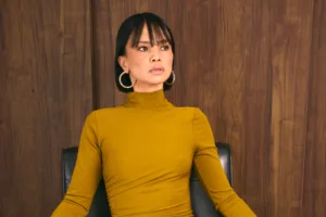 Close-up of a model in a yellow dress with gold hoop earrings against a warm wooden backdrop.