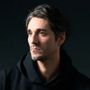 JG, photographe de JG Studio, en sweat à capuche sombre, éclairé en low-key au flash de studio sur fond noir à Shibuya, Tokyo.