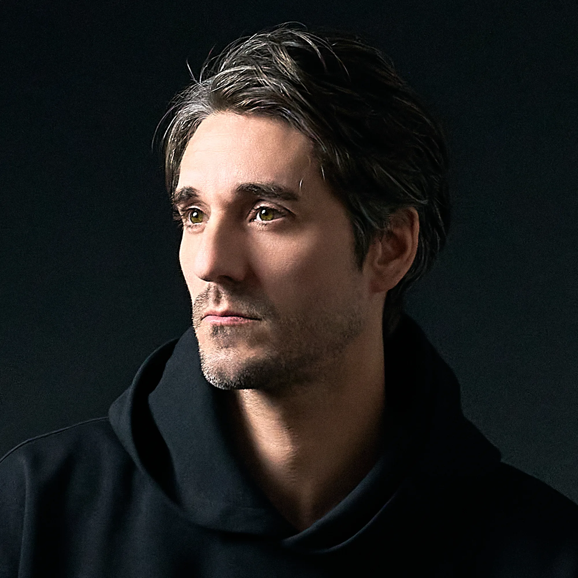 JG, photographer at JG Studio, in a dark hooded top, lit with low-key studio lighting against a black background in Shibuya, Tokyo.