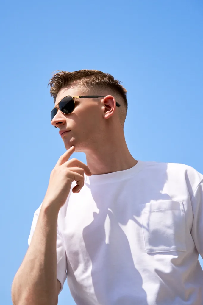 Low-angle outdoor portrait of a man wearing gold-framed sunglasses and a white pocket t-shirt against a clear blue sky, captured during an editorial session in Tokyo by JG.